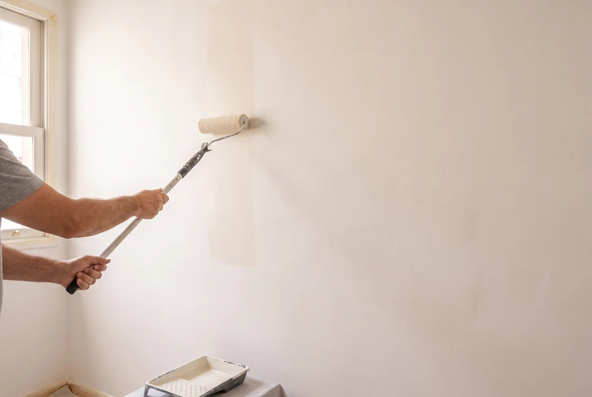 Rolo aplicando tinta clara na parede já preparada, com piso e rodapé protegidos.